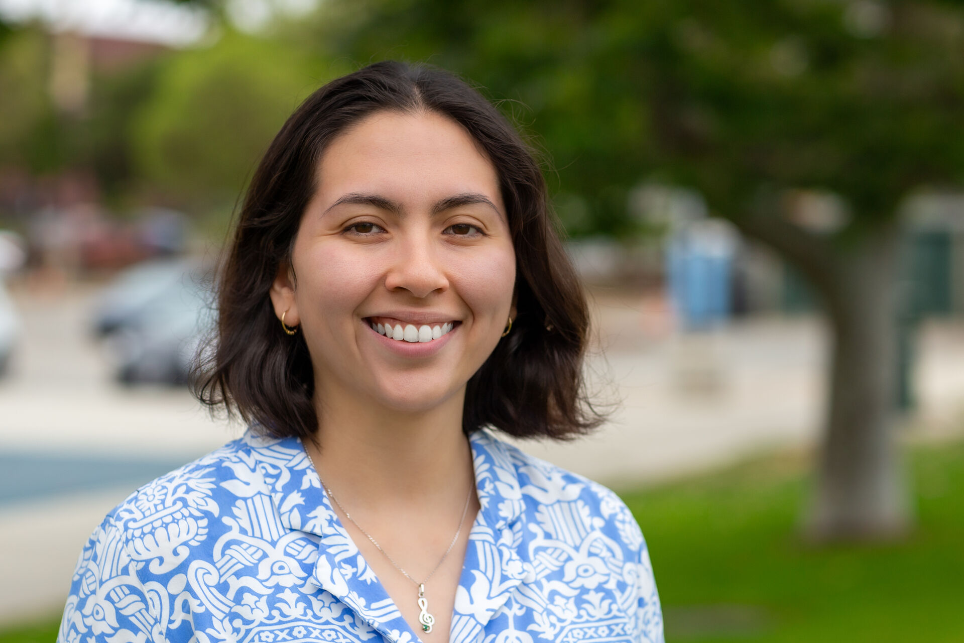 Sonia is standing outside, looking at the camera smiling, with blurred trees in the background. She has shoulder length brown hair and is wearing a blue and white button up shirt + necklace.
