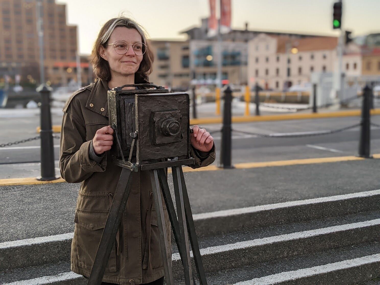 Susannah is standing behind a medium format camera, wearing a khaki knee length jacket and has brown shoulder length hair.