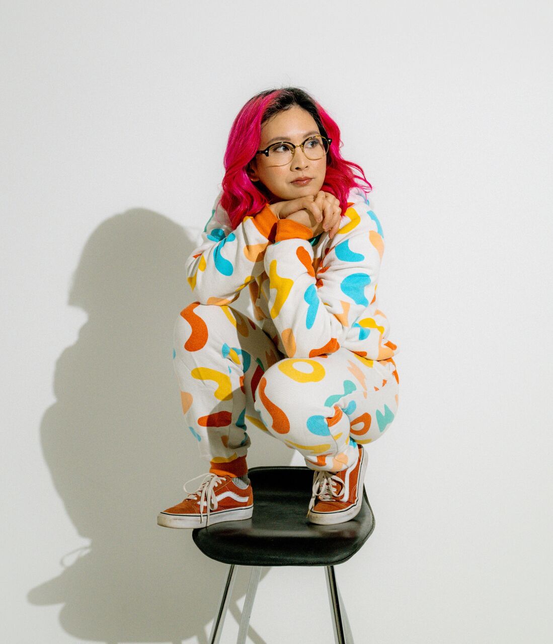 Rad is crouched on a stool with her hands clasped under her gin. She is wearing a white onesie with blue, yellow and red squiggles on it. She has pink hair and is wearing glasses + red shoes.