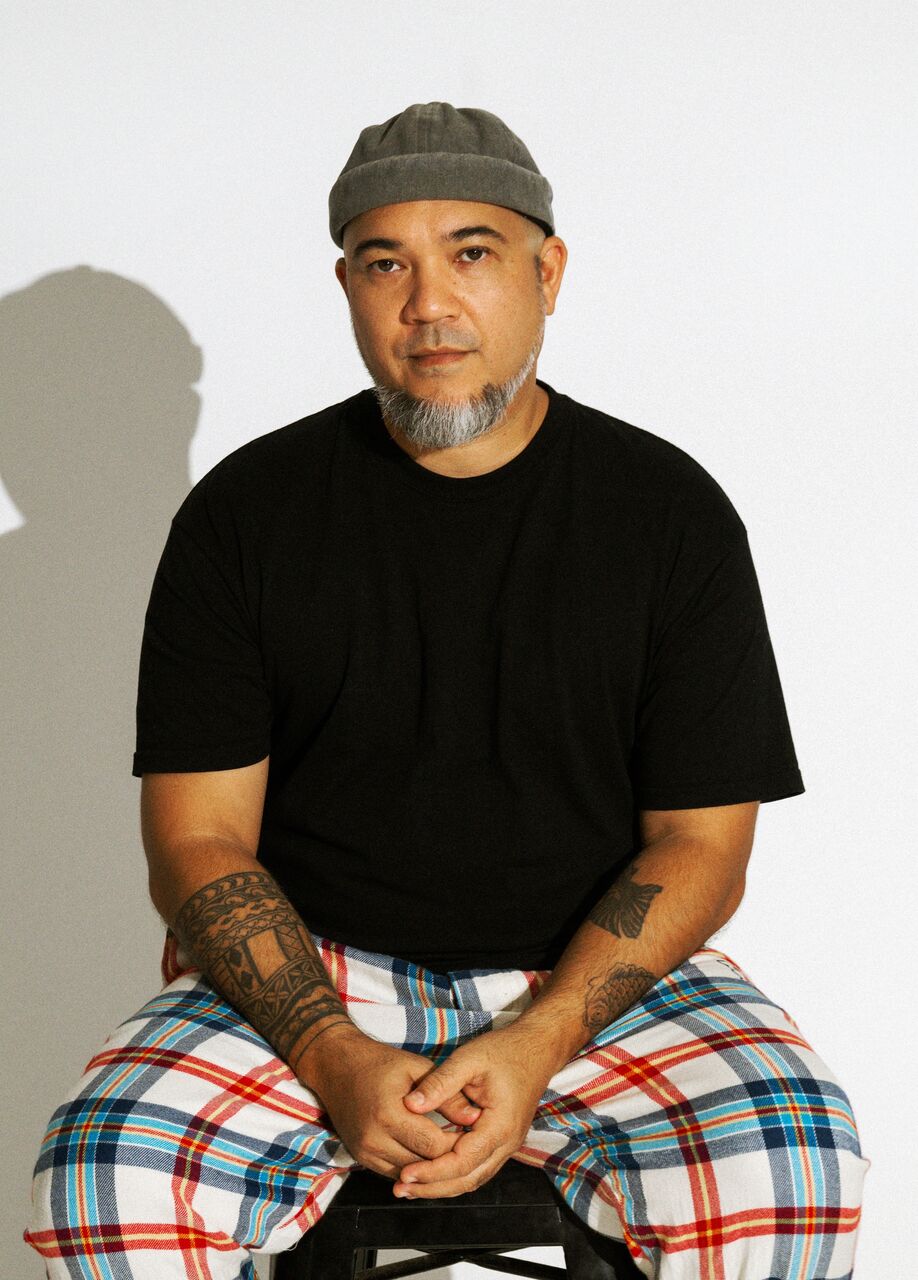 Kuya is sitting on a stool, facing the camera with his hands in his lap. He is wearing a grey beanie, black tshirt and checkered blue, white and red pants.