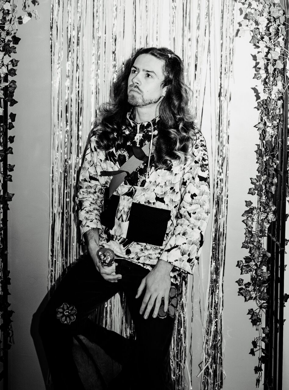 Max is sitting on a stool in front of a glitter streamer backdrop. This is a black and white photograph.
