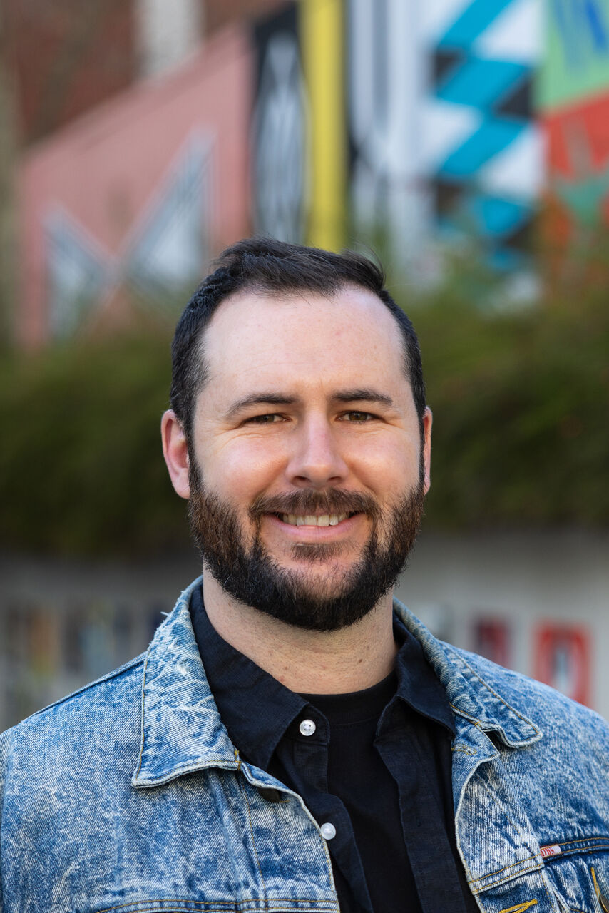 Sam is standing outside, with buildings in the background. He is looking at the camera and has dark hair and a beard. He is wearing a black button up shirt with a denim overshirt.