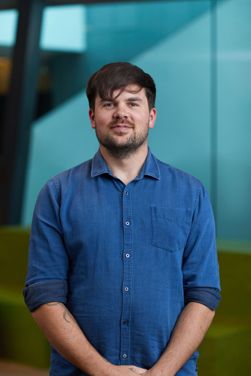 Louie is standing outside with glass buildings behind. He has his hands clasped in front of his body and is looking at the camera. He is wearing a blue button up shirt.