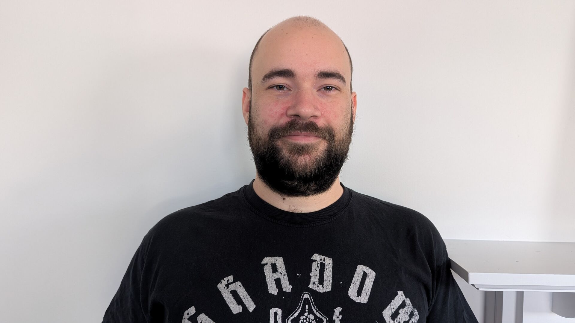 Pascal is standing in front of a white wall, wearing a black graphic t shirt. He has a beard.