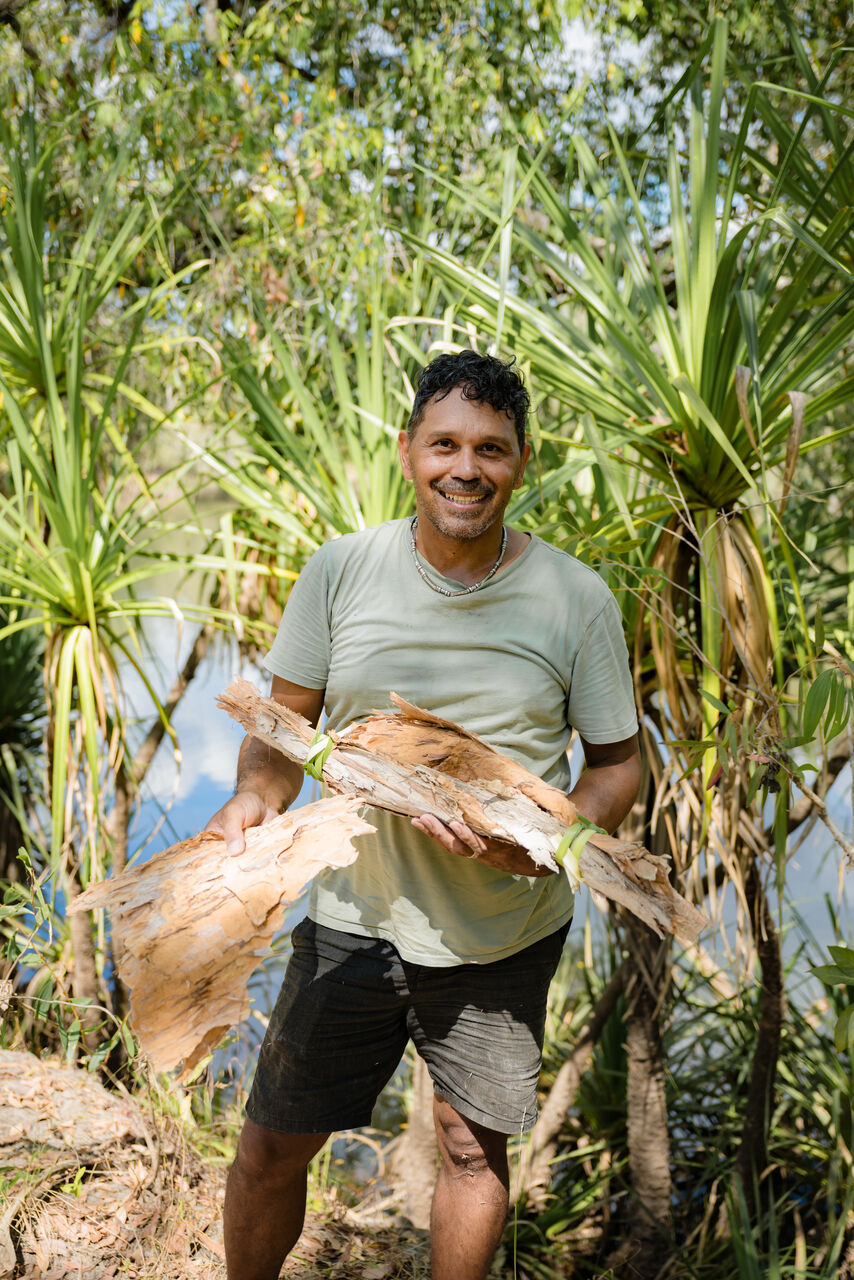 Ben is standing outside with trees and water behind him, holding some tree bark. He is wearing a tshirt and shorts. Smiling and looking at the camera.