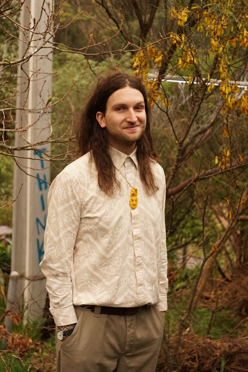 Byron Webb is standing outside, with trees behind him. He has long brown hair and is wearing a button up shirt and a yellow pendant necklace.