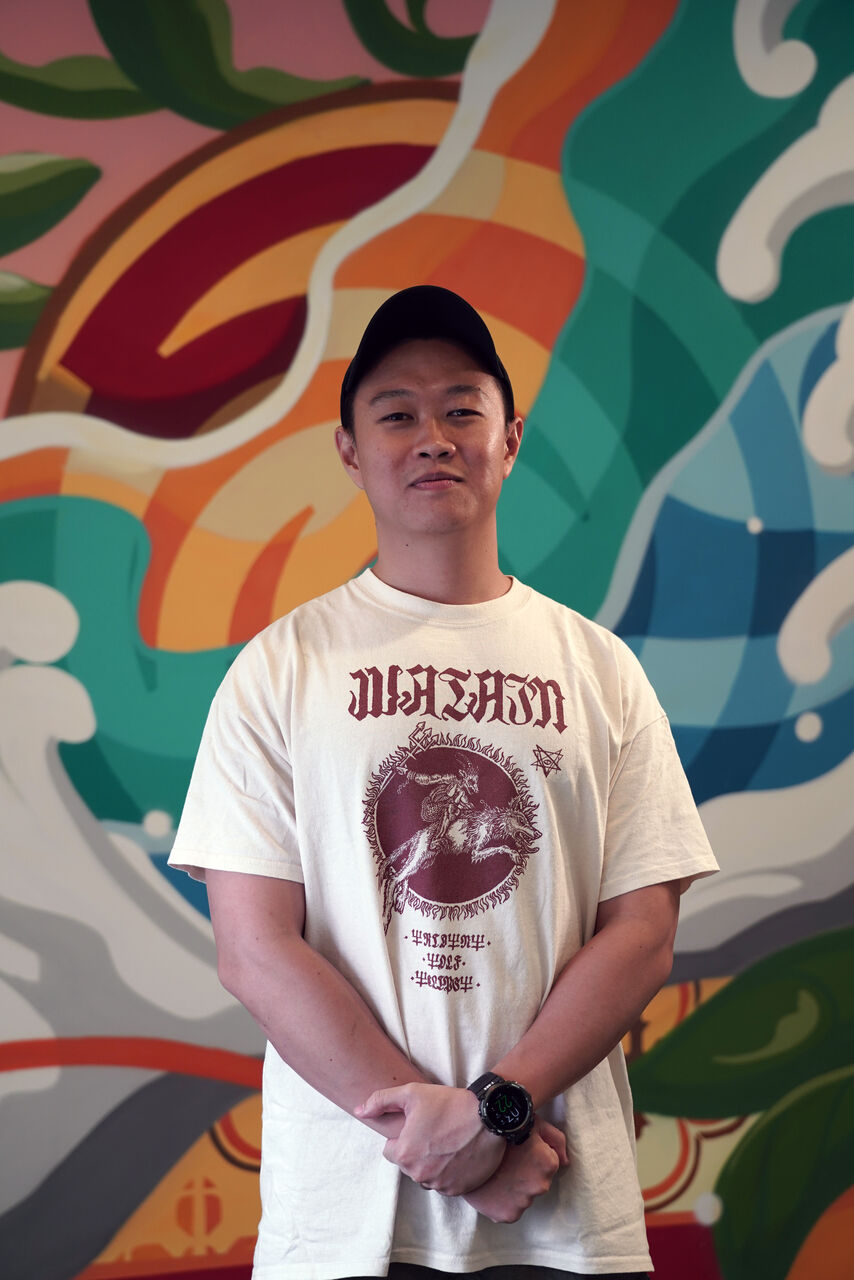 Nathaniel is standing in front of a brightly coloured mural-painted wall. He is wearing a baseball cap and a white tshirt with a maroon logo and writing. He is looking at the camera.