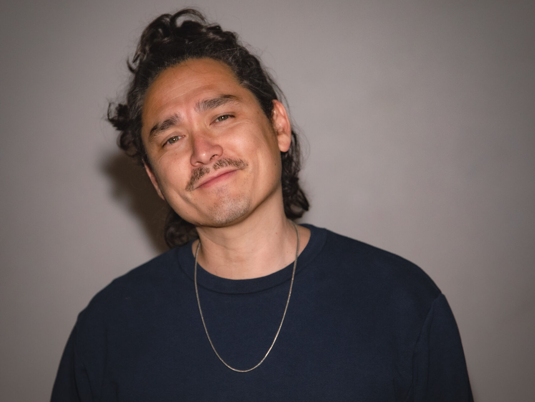 Joel is standing in front of a beige coloured wall. He is dressed all in black and it wearing a long neck chain. He has dark hair and is looking at the camera.