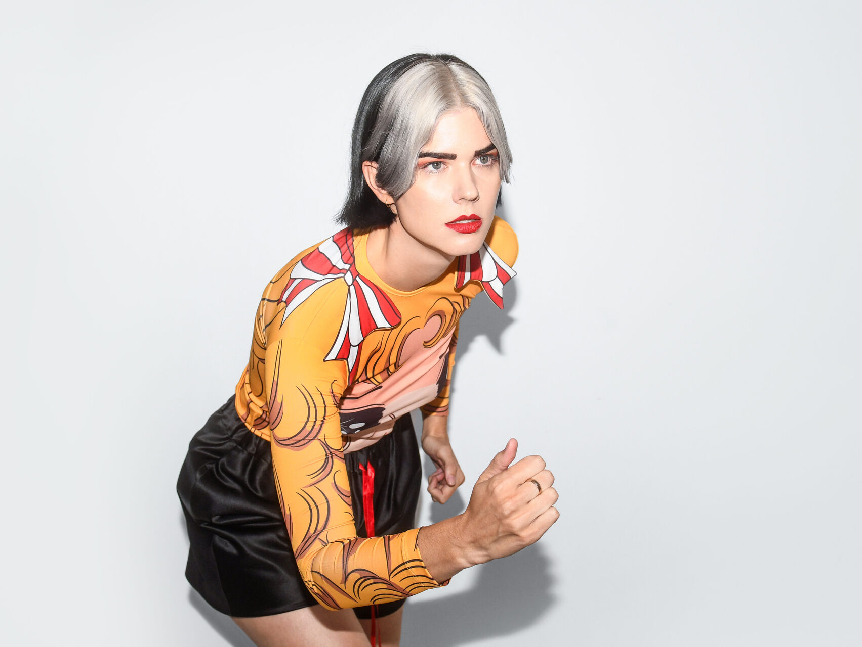 Bec is standing in front of a white backgroup, in a frozen stance like she is running. She has white and black hair and it wearing a graphic long sleeve top with yellow, orange and red colours, and black satin shorts.