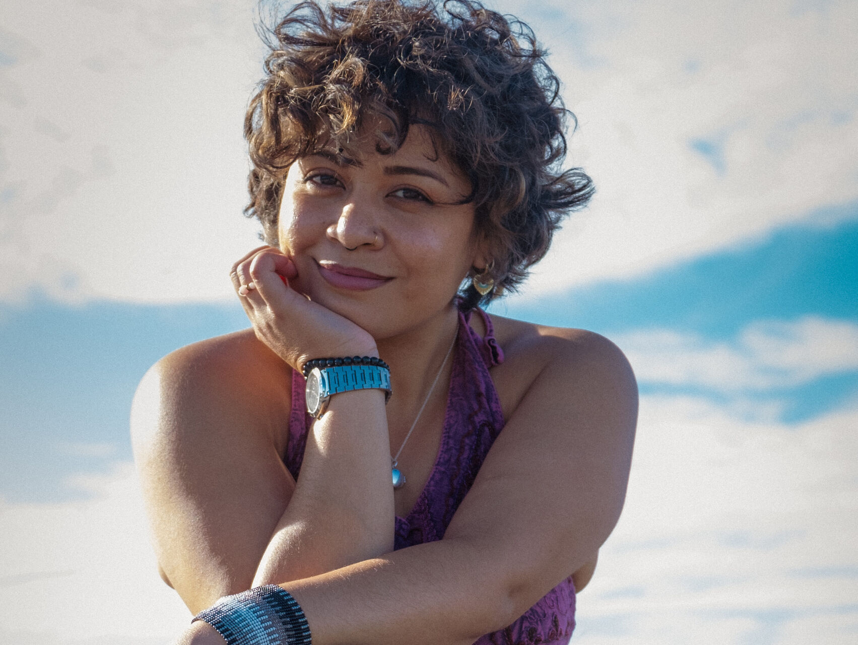 Zeadala is outside, sitting on some rocks. She is wearing black skinny jeans and a purple halter neck top. One of her elbows is resting on her knee & her chin is resting on that hand.