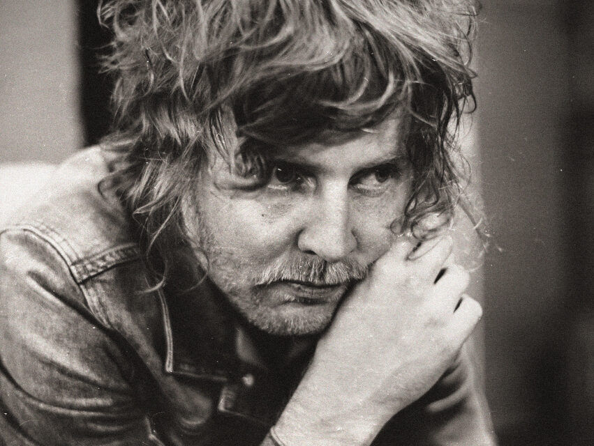 A black & white photo of Kevin who is seated, wearing a denim jacket and leaning his chin onto his hands. He is looking away from the camera.