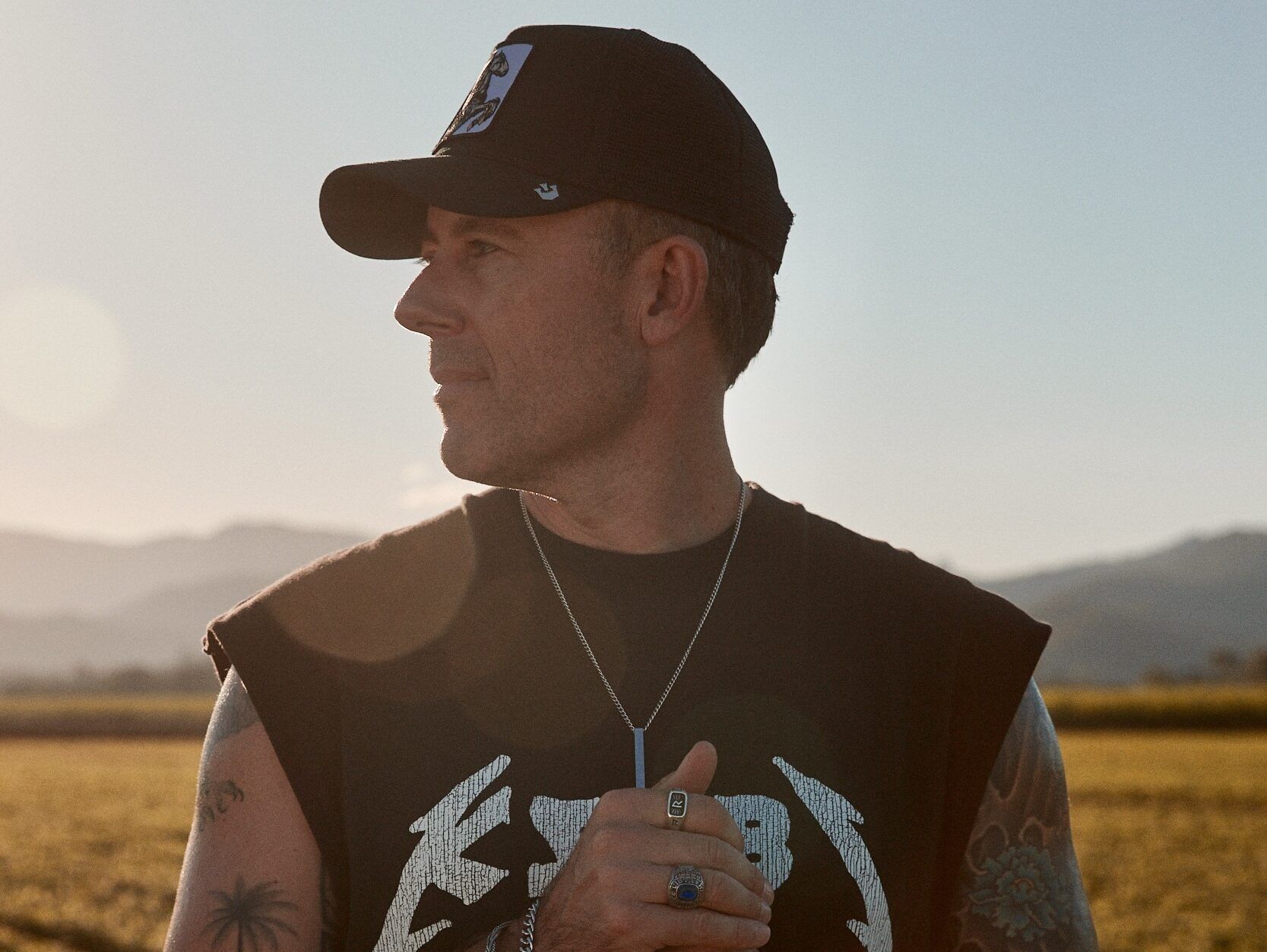 Casey is standing outside in a field, with the sunlight shining behind him. His body is facing the camera and his head is turned. He is wearing a black muscle shirt with white writing and a black cap. His hands are held up in front of his torso. He is wearing rings, a watch and bracelets.