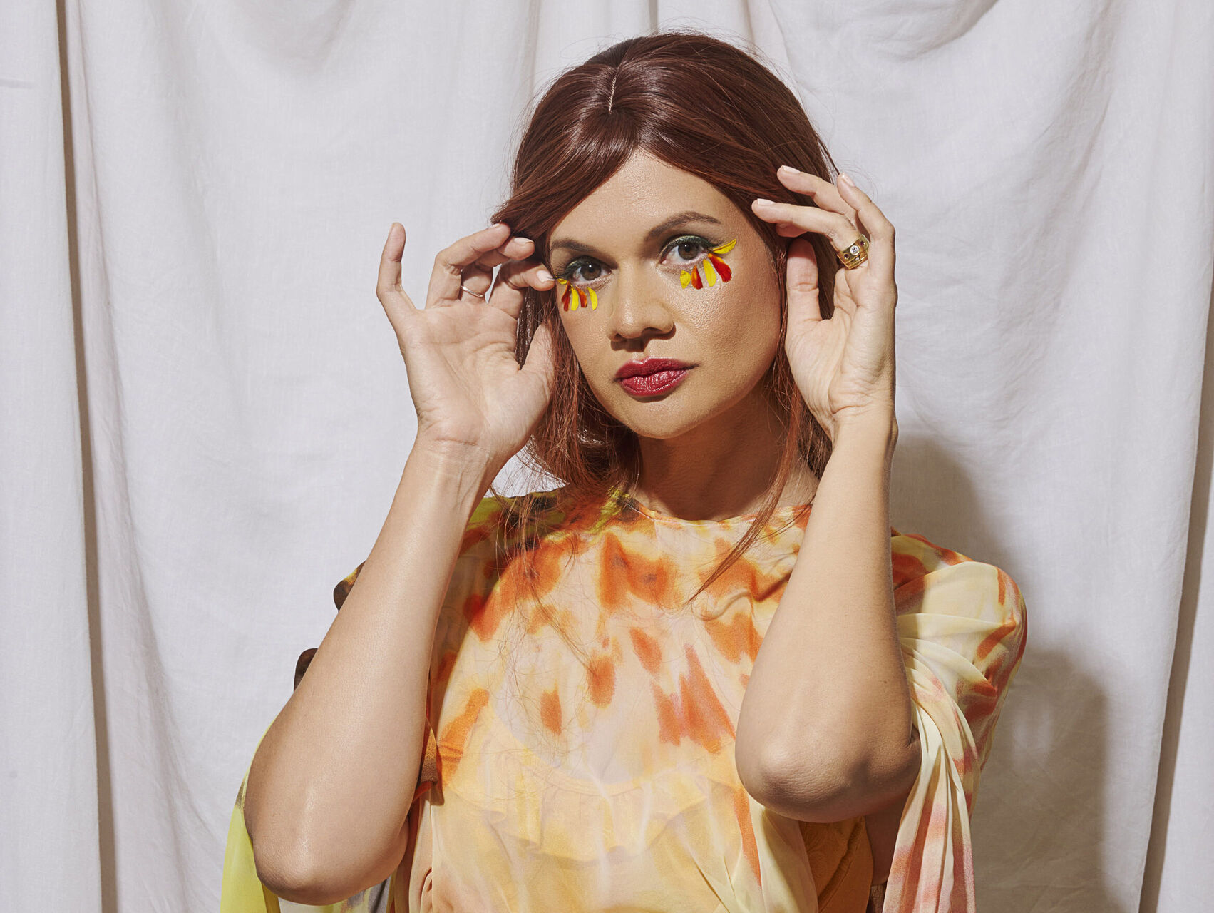 GLVES is standing in front of a white sheet backdrop. She is dressed in colourful clothing of orange, cream, yellow and green. She has small orange & yellow feathers under her eyes. Her hands are held up either side of her face. She has brown hair and is looking at the camera.