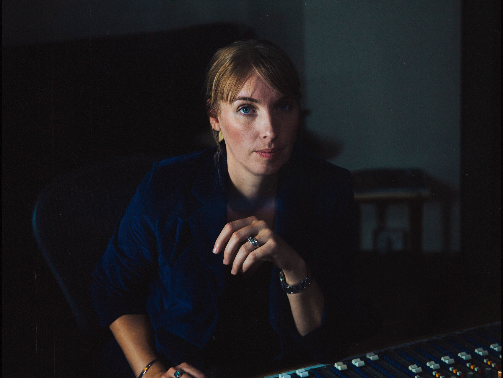 Anna is sitting behind a mixing desk in a studio. She is using the desk with one hand and has her elbow on the table with the other arm. She is wearing navy top and has her hair tied back. She is looking at the camera.