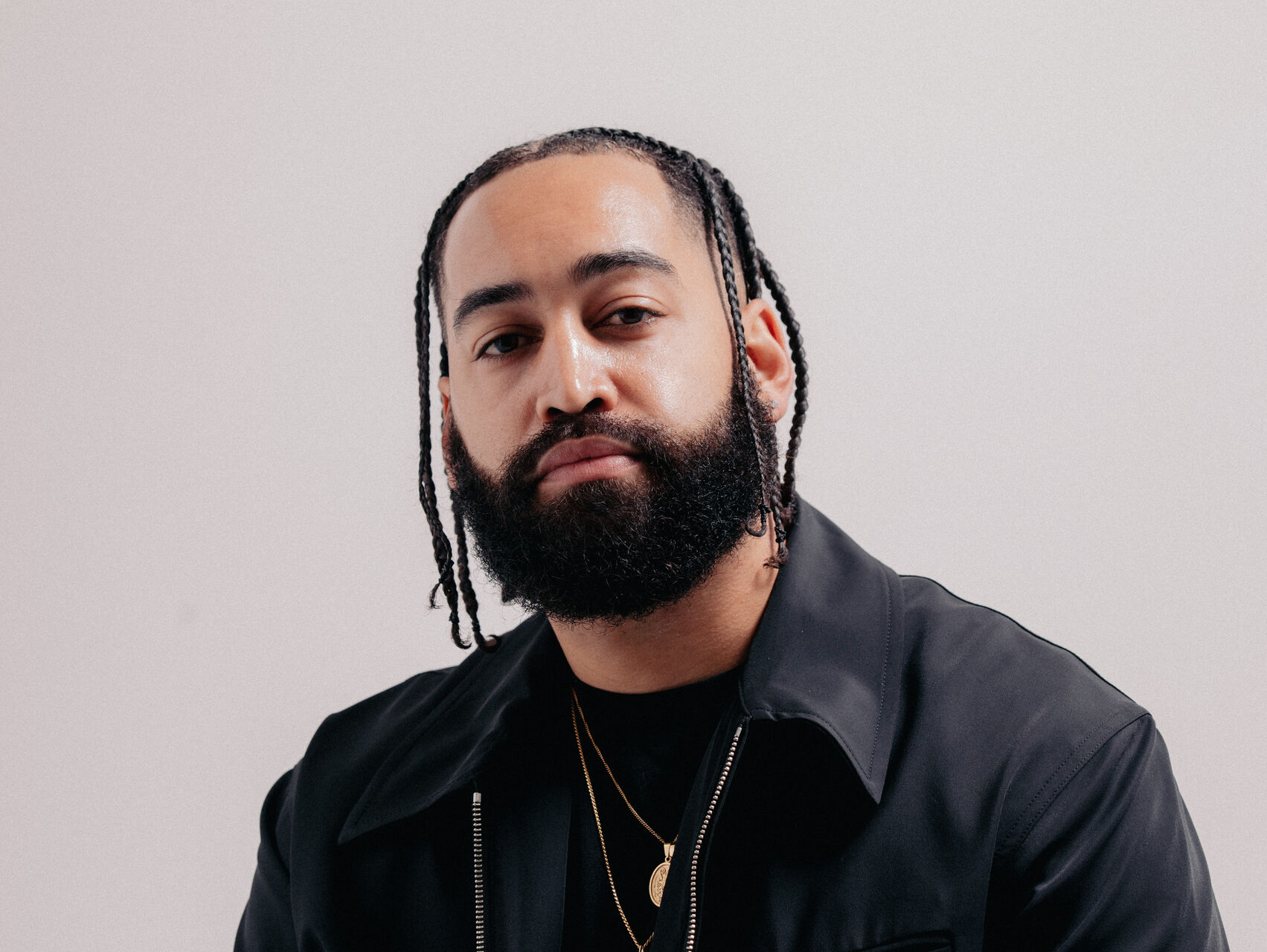 B WISE is standing in front of a beige background. His hair is in plaits and he has a dark beard. He is dressed all in black with a black jacket and a gold chain. He is looking at the camera.