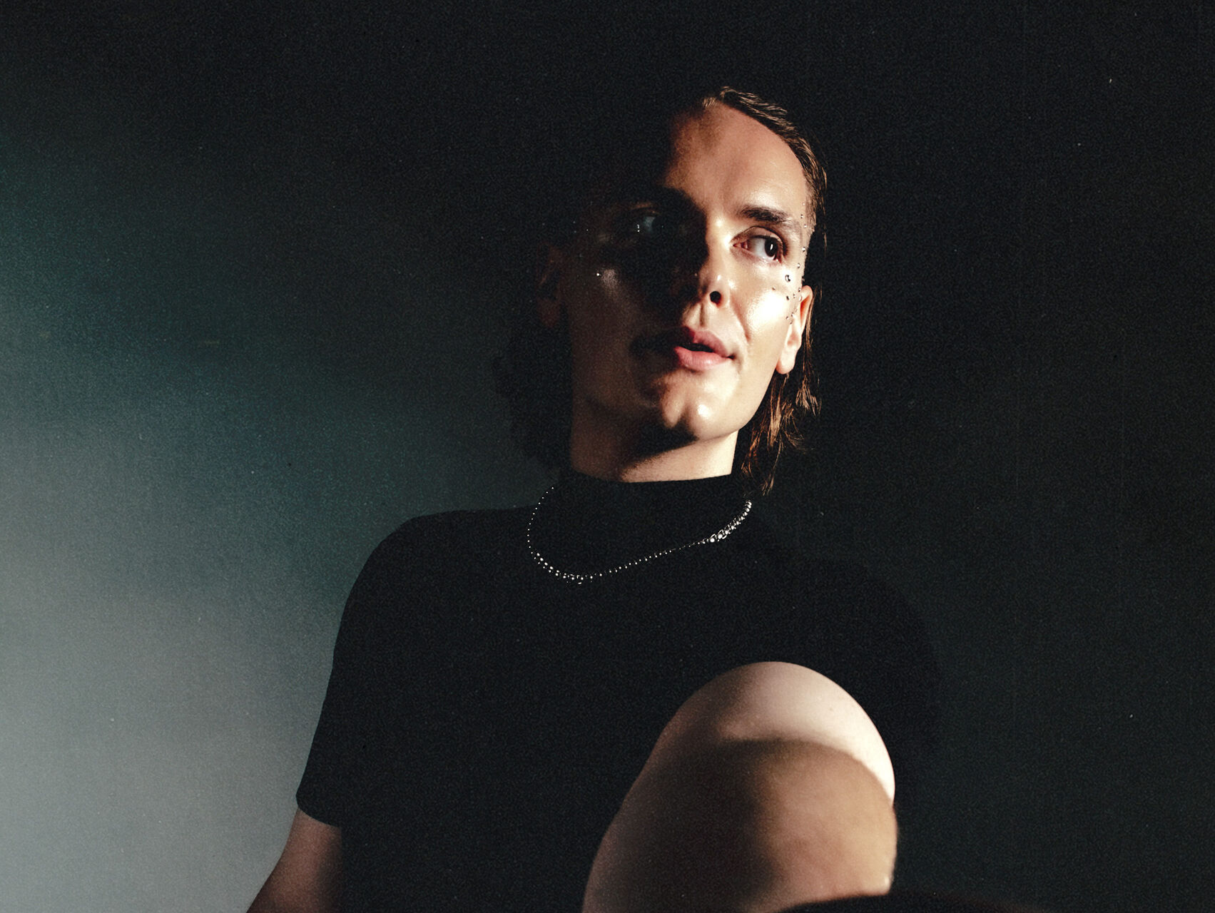 Hylander is standing in the shadows, in front of a dark background. He is dressed all in black and is wearing a necklace. His arm is outstretched towards the camera, palm up. He is looking away from the camera and the light shines onto his face.