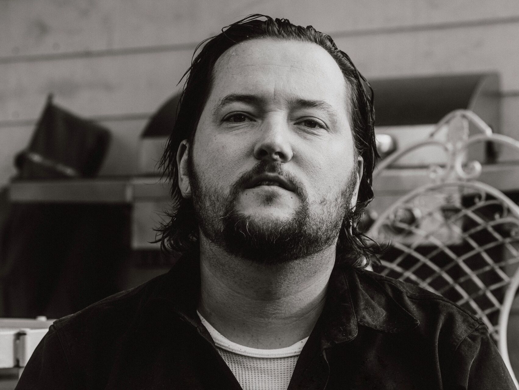 A black & white photo of Wade who is standing in front of what looks like items in a storage shed. He is wearing a black button up shirt with a white tshirt undernearth. His dark hair is slicked back and he has a beard. He is looking at the camera.