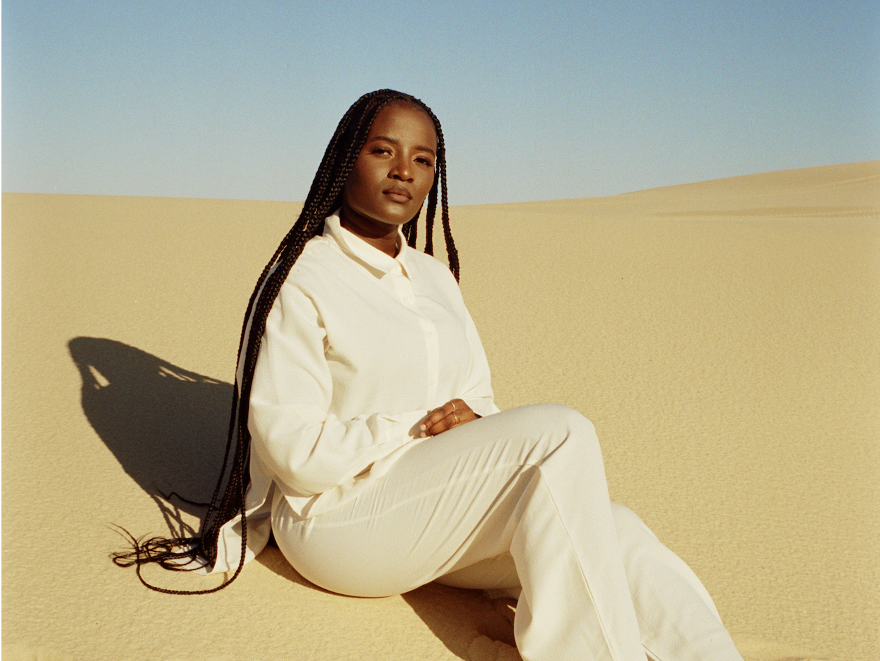 Agum is sitting on a sand dune with blue sky behind her. She is wearing a whirt shirt and white pants, and bare feet. Her hair is long and she is looking at the camera.