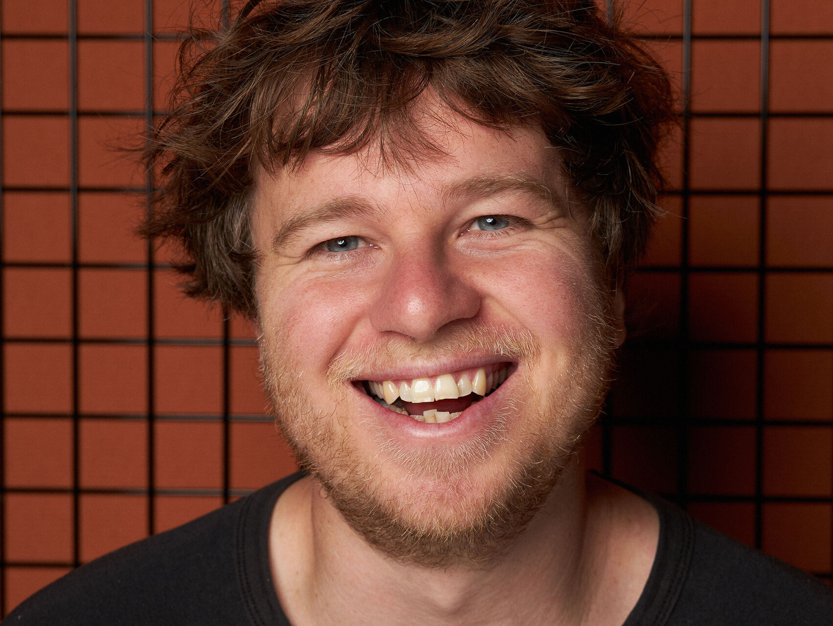 Cody is standing in front of an orange tiled wall, wearing a black & white Bob Dylan tshirt. He is looking at the camera and smiling, almost laughing.