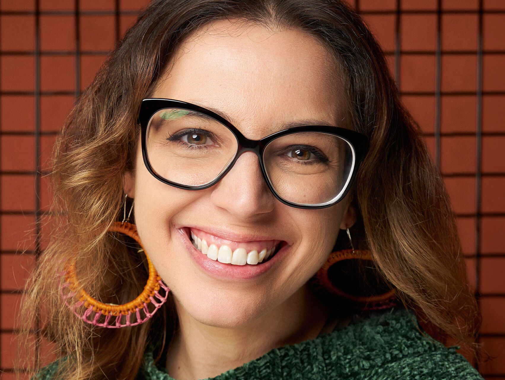 Sophie is standing in front of an orange tiled wall. She is wearing a green jumper, large orange & pink earrings and black framed glasses. She is looking at the camera, smiling.
