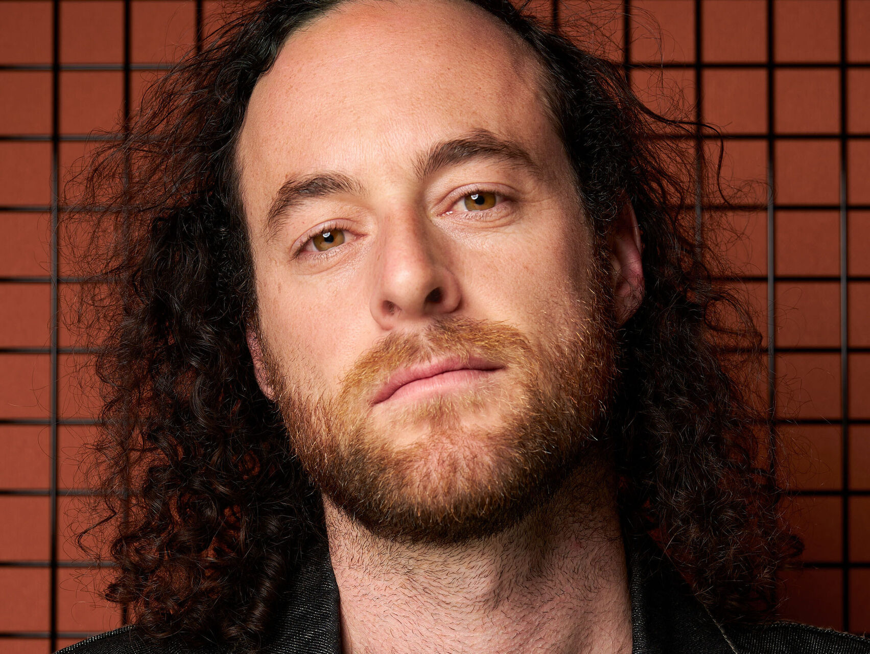 Sam is standing in front of an orange tiled wall. He is wearing a black tshirt and a denim jacket. He has long hair and is looking at the camera.