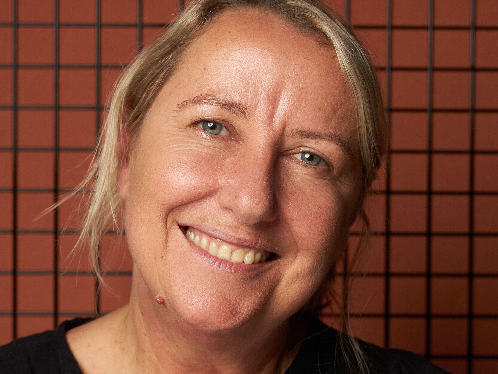 Head shot of Ali who is standing in front of an orange tiled wall. She is wearing a black top and has blonde hair. She is looking at the camera, smiling.