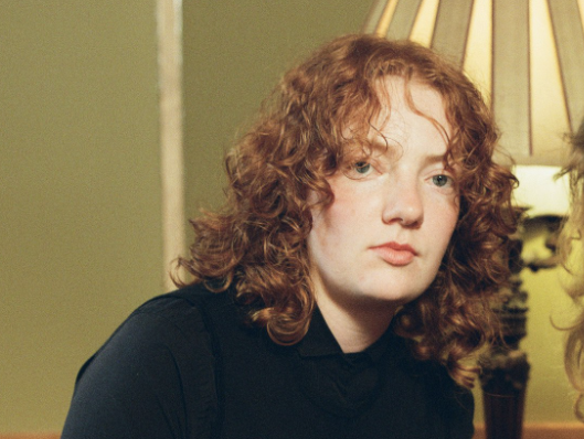 Heide is sitting in a lounge room setting with a lamp behind them. They are dressed in all black and have red curly shoulder length hair. They are looking at the camera.