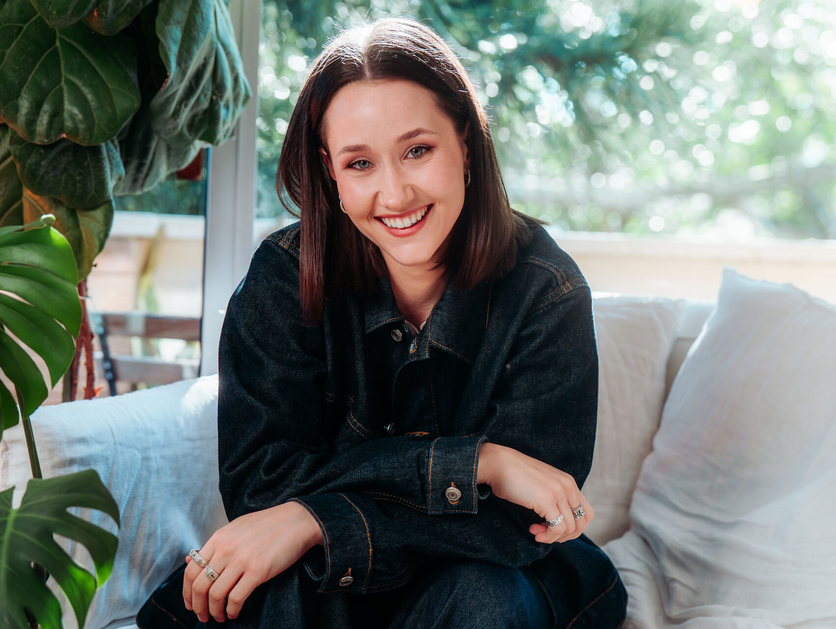 Ash is sitting on a couch in front of a glass window and you can see a balcony and trees outside. There is a monstera plant next to her. She is wearing all black and is looking at the camera smiling. She has long dark hair.