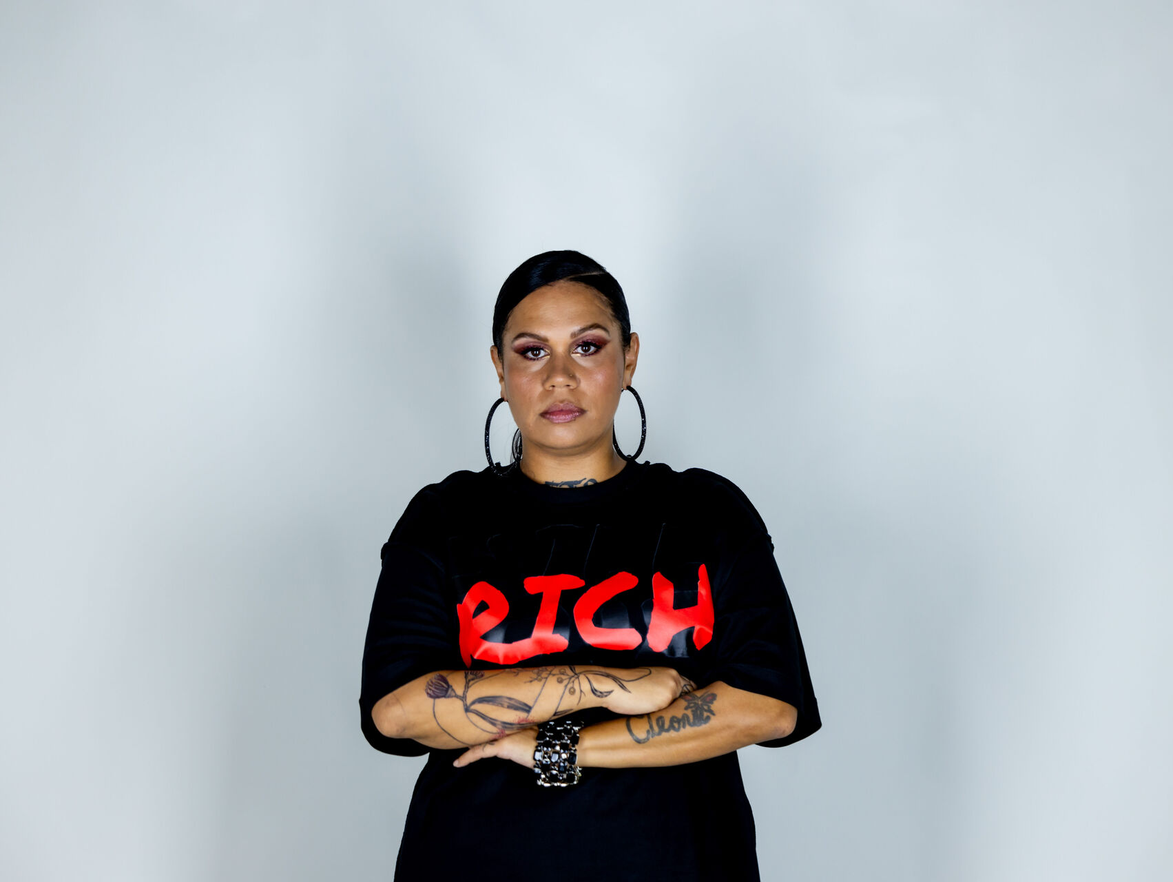 BARKAA is standing in front of a white backdrop with her arms crossed. She is wearing a black tshirt with the word RICH written in capital letters in red. She has large hoop earrings, dark hair and is looking directly into the camera.