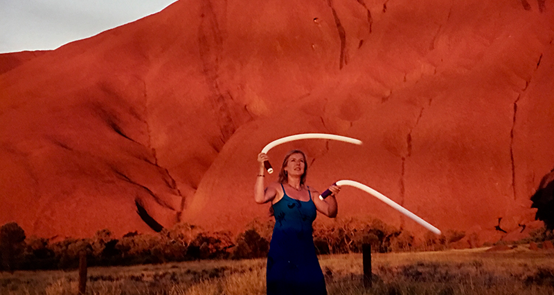 Sarah Hopkins stands in front of Uluru in Australia, holding two harmonic whirlies, a wind instrument she invented